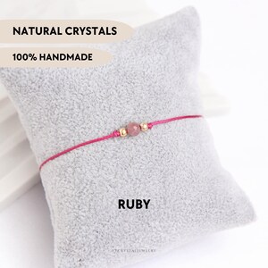 Op de afbeelding: Een delicate armband met een enkele gefacetteerde robijnkraal geflankeerd door twee kleine goudkleurige kralen aan een dun roze koord. De tekst NATURAL CRYSTALS, 100% HANDMADE en RUBY is zichtbaar. Het ontwerp is eenvoudig en minimalistisch.