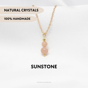 Op de afbeelding: Een goudkleurige ketting met een hanger van twee ronde, perzikkleurige zonnesteenkralen, gescheiden door een kleine goudkleurige kraal. De tekst NATURAL CRYSTALS, 100% HANDMADE, SUNSTONE en YPCRYSTALJEWELRY is zichtbaar op de afbeelding.