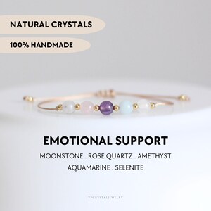 Puede incluir: Una pulsera delicada con cinco cuentas redondas de piedra luna, cuarzo rosa, amatista, aguamarina y selenita, separadas por pequeños espaciadores de metal dorado en un cordón fino. Texto: NATURAL CRYSTALS, 100% HANDMADE, EMOTIONAL SUPPORT.