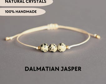 May include: A bracelet featuring three round Dalmatian jasper beads with black spots on a cream-colored cord, accented by small gold-toned metal spacers. The image includes the text NATURAL CRYSTALS, 100% HANDMADE, and DALMATIAN JASPER.