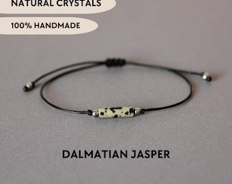 May include: A bracelet featuring a cylindrical Dalmatian jasper bead with black spots on a cream background, flanked by two small silver-toned metal beads on a thin black cord. Text reads: NATURAL CRYSTALS, 100% HANDMADE, DALMATIAN JASPER.
