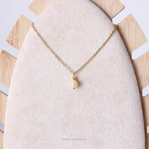 Puede incluir: Un collar de cadena dorada fina con un colgante de una peque&ntilde;a cuenta redonda de color crema p&aacute;lido. El collar se presenta sobre un soporte texturizado de color claro en forma de gota. El texto YFCRYSTALJEWELRY est&aacute; impreso en la parte inferior.