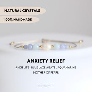 Op de afbeelding: Een armband gemaakt van natuurlijke kristallen, waaronder angeliet, blauwe kant agaat, aquamarijn en parelmoer. De armband ligt op een witte achtergrond met de tekst "NATURAL CRYSTALS" en "100% HANDMADE" boven de armband en de tekst "ANXIETY RELIEF" en "ANGELITE. BLUE LACE AGATE. AQUAMARINE MOTHER OF PEARL" onder de armband.