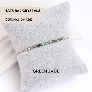 Op de afbeelding: Een delicate armband met een dun beige koord, geregen met afwisselend kleine cilindrische groene jade kralen en piepkleine zilverkleurige metalen bolletjes. Het ontwerp is eenvoudig en lineair en toont natuurlijke groene stenen.