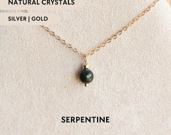 Può includere: Una collana a catena color oro con un ciondolo composto da una singola perla rotonda in serpentino verde scuro. Il testo sull'immagine recita NATURAL CRYSTALS, SILVER | GOLD e SERPENTINE. Il gioiello è esposto su un materiale chiaro e testurizzato.