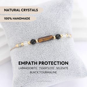 May include: A beaded bracelet featuring a central rectangular tiger's eye bead, flanked by black tourmaline, labradorite, and selenite beads, accented with small gold-toned metal spacers on a thin cord. Text reads NATURAL CRYSTALS 100% HANDMADE.