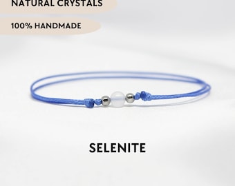 Könnte beinhalten: Ein blaues Schnurarmband mit einer zentralen weißen Selenitperle, flankiert von zwei kleinen metallisch silbernen Perlen. Das Bild enthält den Text NATURAL CRYSTALS, 100% HANDMADE, SELENITE und YPCRYSTALJEWELRY. Das Design verwendet eine einfache Knotenschnur.