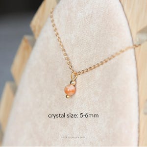 Puede incluir: Un collar de cadena dorada con un colgante de una sola cuenta de cristal redonda de color melocot&oacute;n con peque&ntilde;os detalles dorados. El texto crystal size: 5-6mm aparece debajo del colgante y YPCRYSTALJEWELRY en la parte inferior.