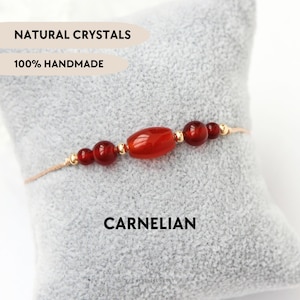 Può includere: Un braccialetto con una perla centrale ovale in corniola affiancata da perle rotonde più piccole in corniola e minuscoli distanziatori dorati su un cordino sottile. Il testo riporta NATURAL CRYSTALS, 100% HANDMADE, CARNELIAN.