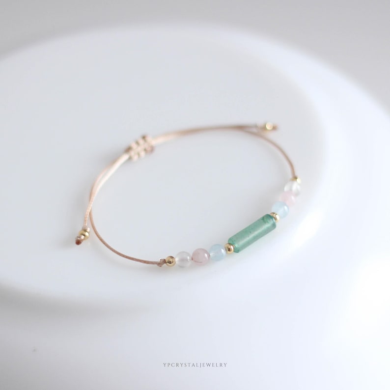 May include: Adjustable bracelet featuring a light beige cord and a series of colorful beads. The bracelet includes a green rectangular bead, pink and clear round beads, and small gold-colored accents. The brand name "YPCYSTALJEWELRY" is printed at the bottom.