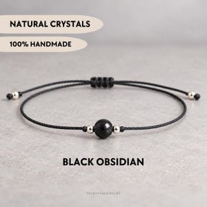 Può includere: Un bracciale in cordino nero con una perla centrale in ossidiana nera, affiancata da due piccole perle metalliche color argento. La chiusura regolabile presenta un design in cordino intrecciato. Testo: NATURAL CRYSTALS, 100% HANDMADE, BLACK OBSIDIAN.