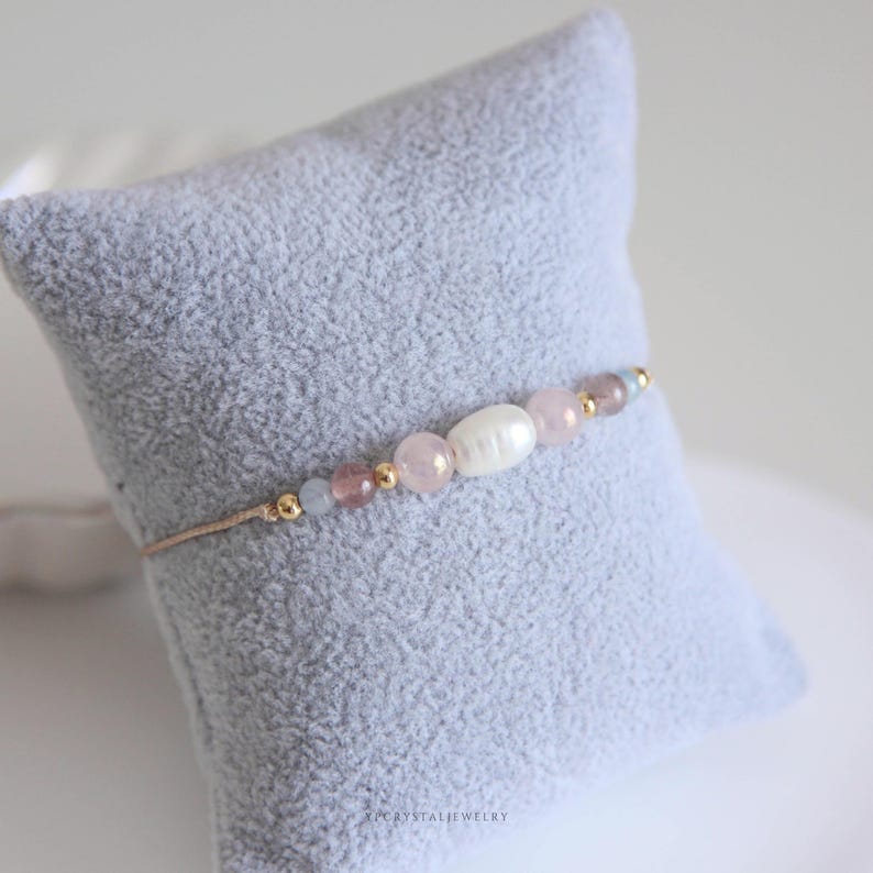 May include: A delicate bracelet featuring a central white oval pearl flanked by round pink and light blue gemstone beads, interspersed with small gold-toned metal spheres on a thin cord. The text YICRYSTALJEWELRY is visible at the bottom.