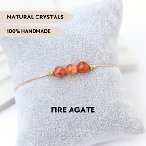 Puede incluir: Un brazalete hecho a mano con tres cuentas de ágata de fuego naranja y detalles dorados. El brazalete está ensartado en un cordón beige y se muestra sobre un cojín gris. El texto de la imagen dice: "Natural Crystals", "100% Handmade" y "Fire Agate".