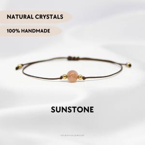 Può includere: Un braccialetto delicato con una singola perla rotonda color pesca in pietra del sole, affiancata da due piccole perline metalliche dorate su un sottile cordino marrone. Il testo NATURAL CRYSTALS, 100% HANDMADE e SUNSTONE è visibile.