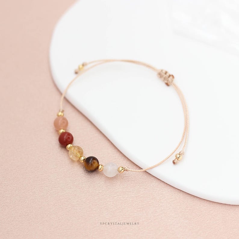 May include: A bracelet with a series of colourful gemstones, including pink, red, yellow, brown, and white. The beads are strung on a beige cord with gold-coloured accents. The bracelet is displayed on a white surface with a pink background.