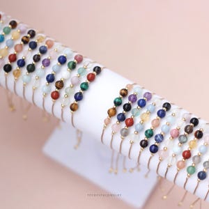 May include: A collection of adjustable bracelets featuring various colorful natural stone beads and small gold-toned metal spacers. Each bracelet displays a unique sequence of polished spherical gemstones in shades of blue, green, red, yellow, and purple. The text YPCRYSTALJEWELRY is visible at the bottom.