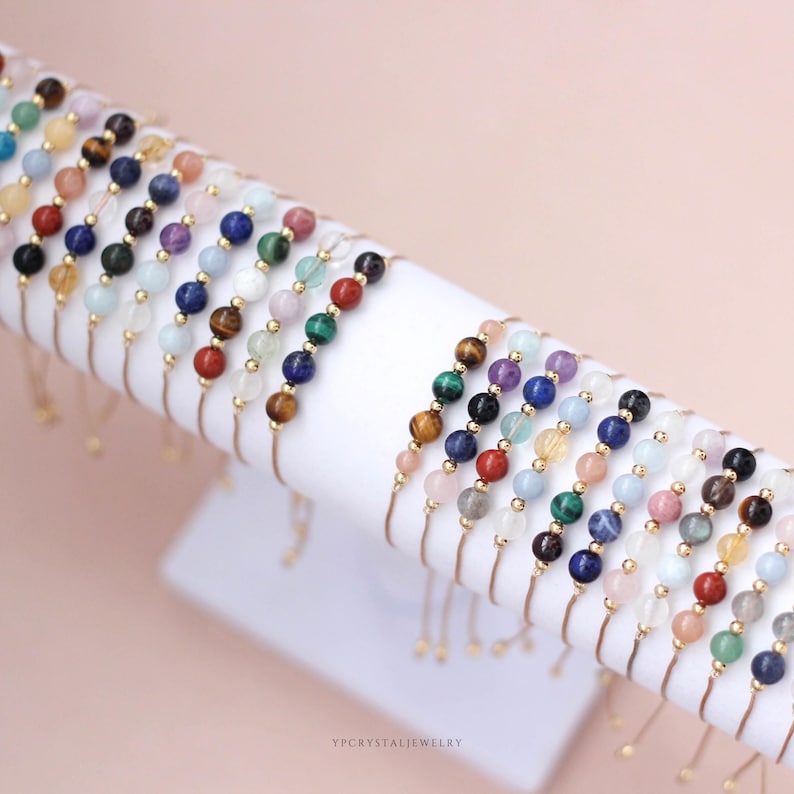 May include: A collection of adjustable bracelets featuring various colorful natural stone beads and small gold-toned metal spacers. Each bracelet displays a unique sequence of polished spherical gemstones in shades of blue, green, red, yellow, and purple. The text YPCRYSTALJEWELRY is visible at the bottom.