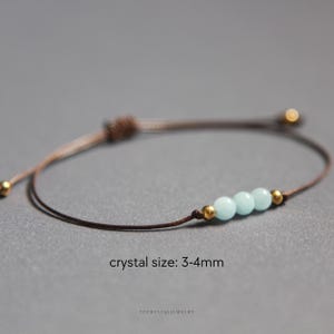 K&ouml;nnte beinhalten: Ein braunes Kordelarmband mit drei hellblauen Perlen, flankiert von zwei kleinen goldfarbenen Metallperlen. Das Armband hat einen verstellbaren Schiebeknotenverschluss und enth&auml;lt den Text crystal size: 3-4mm und ypcrystaljewelry.