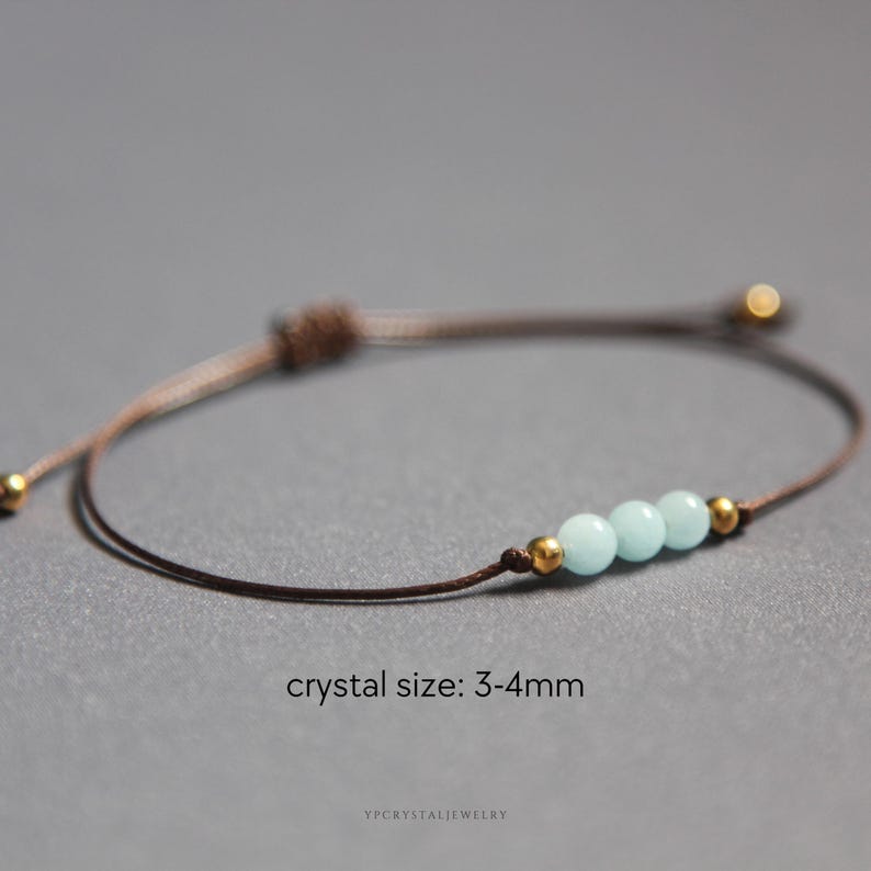 K&ouml;nnte beinhalten: Ein braunes Kordelarmband mit drei hellblauen Perlen, flankiert von zwei kleinen goldfarbenen Metallperlen. Das Armband hat einen verstellbaren Schiebeknotenverschluss und enth&auml;lt den Text crystal size: 3-4mm und ypcrystaljewelry.