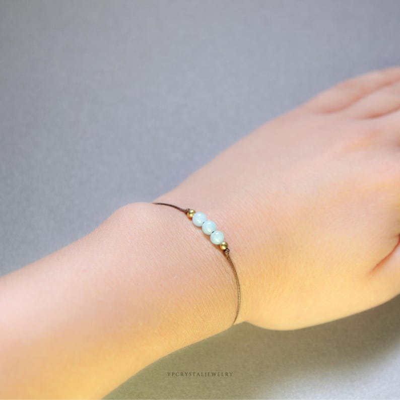 K&ouml;nnte beinhalten: Ein zartes Armband mit drei hellblauen, kugelf&ouml;rmigen Perlen, die zwischen zwei kleinen metallisch goldfarbenen Perlen auf einer d&uuml;nnen braunen Kordel angeordnet sind. Der Text YPCRYSTALJEWELRY steht in kleinen schwarzen Buchstaben unten.