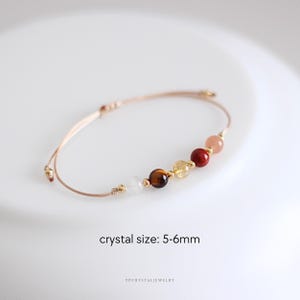 May include: A delicate bracelet with a series of colourful crystal beads, each approximately 5-6mm in diameter. The beads include white, brown, yellow, red, and pink hues, strung on a thin, beige cord with gold accents. The text "crystal size: 5-6mm" is visible.
