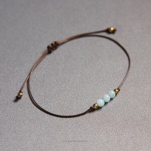 K&ouml;nnte beinhalten: Ein Armband aus brauner Kordel mit drei hellblauen runden Perlen, die von zwei kleinen goldfarbenen Metallperlen flankiert werden. Das Armband verf&uuml;gt &uuml;ber einen verstellbaren Schiebeknotenverschluss mit zwei weiteren Goldperlen an den Enden.