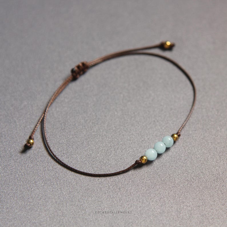 K&ouml;nnte beinhalten: Ein Armband aus brauner Kordel mit drei hellblauen runden Perlen, die von zwei kleinen goldfarbenen Metallperlen flankiert werden. Das Armband verf&uuml;gt &uuml;ber einen verstellbaren Schiebeknotenverschluss mit zwei weiteren Goldperlen an den Enden.