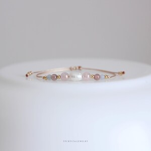 May include: A delicate adjustable bracelet featuring a central white baroque pearl flanked by soft pink and light blue gemstone beads, interspersed with small gold-toned metallic spheres on a thin, light-colored cord with sliding knots.