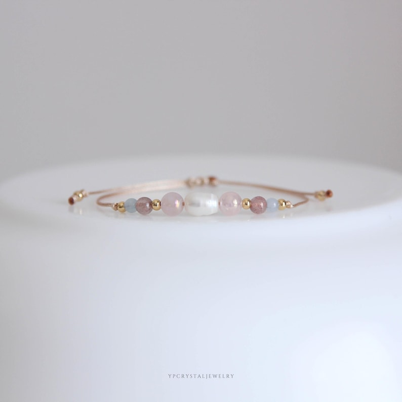 May include: A delicate adjustable bracelet featuring a central white baroque pearl flanked by soft pink and light blue gemstone beads, interspersed with small gold-toned metallic spheres on a thin, light-colored cord with sliding knots.