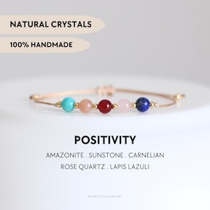 以下が含まれることがあります： アマゾナイト、サンストーン、カーネリアン、ローズクォーツ、ラピスラズリなど、天然のクリスタルで作られたブレスレット。ブレスレットは薄い茶色の紐に、各クリスタル間に金色のビーズが施されています。ブレスレットの下には「POSITIVITY」という文字、画像の上部には「NATURAL CRYSTALS 100% HANDMADE」という文字が書かれています。