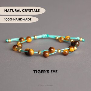 Könnte beinhalten: Ein Armband mit braunen Tigerauge-Perlen und kleinen goldfarbenen Metallperlen auf einem helltürkisen Band. Das Bild enthält den Text NATURAL CRYSTALS, 100% HANDMADE und TIGER'S EYE. Das Design verfügt über einen verstellbaren Schiebeknoten.