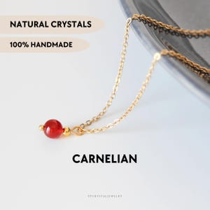 Puede incluir: Un collar de cadena en tono dorado con una cuenta redonda de cornalina roja pulida, acentuada por pequeñas cuentas de metal en tono dorado. La imagen incluye el texto NATURAL CRYSTALS, 100% HANDMADE, CARNELIAN y YPCRYSTALJEWELRY.