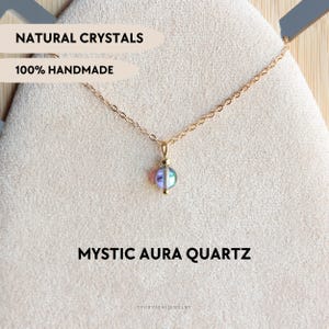 Puede incluir: Un collar de cadena dorada con un colgante de cuenta redonda de cuarzo aura místico iridiscente. El texto NATURAL CRYSTALS, 100% HANDMADE y MYSTIC AURA QUARTZ es visible. La pieza de joyería se muestra sobre una tela texturizada neutra.