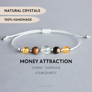 Peut inclure: Un bracelet sur cordon blanc avec cinq perles : deux citrines jaunes, deux œils-de-tigre marron et une perle de quartz clair au centre, séparées par des petites entretoises argentées. Texte : NATURAL CRYSTALS, 100% HANDMADE, MONEY ATTRACTION, CITRINE, TIGER'S EYE, CLEAR QUARTZ.