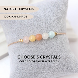 Può includere: Un braccialetto con cinque perline rotonde in cristallo naturale multicolore separate da piccoli distanziatori in metallo dorato su un cordino beige. Il testo recita: NATURAL CRYSTALS, 100% HANDMADE, CHOOSE 5 CRYSTALS CORD COLOR AND SPACER BEADS.