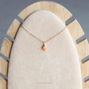 Puede incluir: Un collar de cadena dorada delicada con un colgante de piedra preciosa peque&ntilde;a, redonda y de color melocot&oacute;n centrado en la cadena. El colgante est&aacute; acentuado por una peque&ntilde;a cuenta dorada justo encima de la piedra. El texto YPCRYSTALJEWELRY aparece abajo.