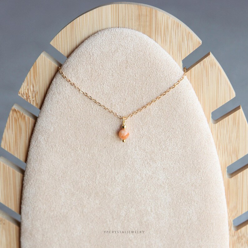 Puede incluir: Un collar de cadena dorada delicada con un colgante de piedra preciosa peque&ntilde;a, redonda y de color melocot&oacute;n centrado en la cadena. El colgante est&aacute; acentuado por una peque&ntilde;a cuenta dorada justo encima de la piedra. El texto YPCRYSTALJEWELRY aparece abajo.