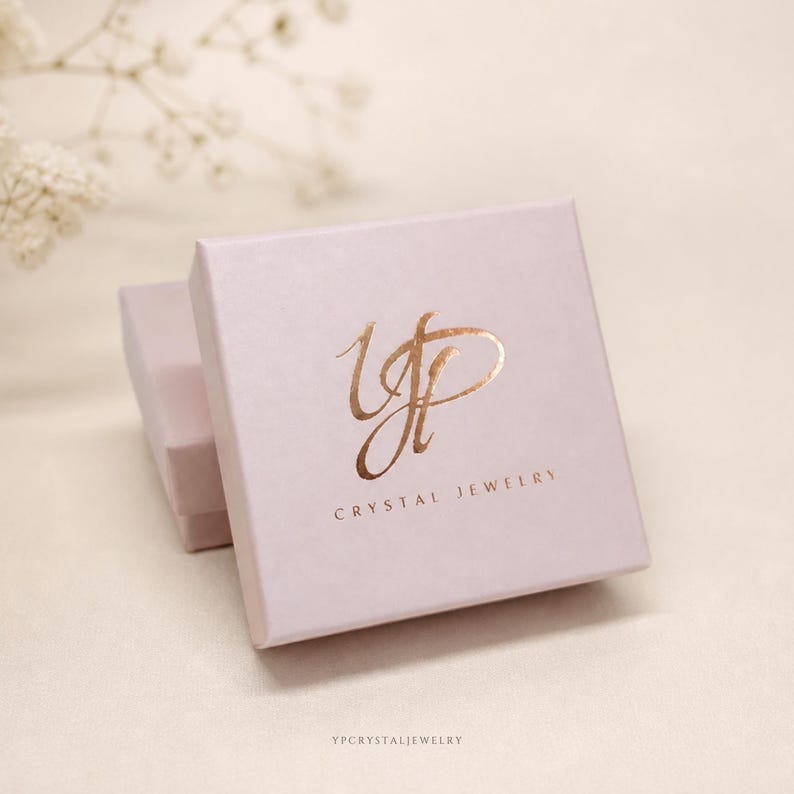 Puede incluir: Una caja de joyer&iacute;a cuadrada de color rosa claro con un logotipo de l&aacute;mina de oro rosa met&aacute;lico en la tapa. El logotipo muestra letras cursivas estilizadas y el texto CRYSTAL JEWELRY impreso en una fuente sans-serif debajo del dise&ntilde;o.