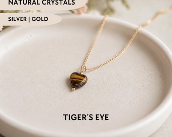 May include: A gold-toned chain necklace featuring a small, heart-shaped tiger's eye pendant. The text NATURAL CRYSTALS, SILVER | GOLD, and TIGER'S EYE is visible. The jewelry piece is displayed on a smooth, off-white circular dish.