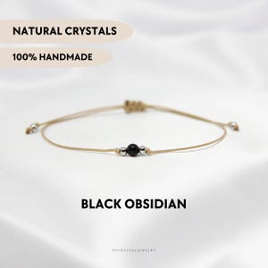 Può includere: Un braccialetto delicato con una singola perla di ossidiana nera affiancata da due piccole perline metalliche color argento su un sottile cordino beige. Il testo NATURAL CRYSTALS, 100% HANDMADE, BLACK OBSIDIAN e YPCRYSTALJEWELRY è visibile.
