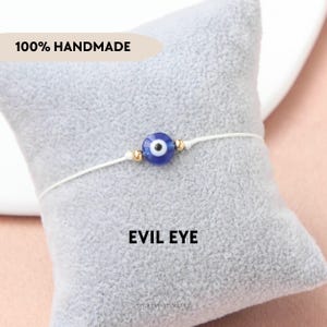 Può includere: Un braccialetto fatto a mano con un ciondolo a occhio blu e bianco, perline dorate e un cordino bianco. Il testo "100% HANDMADE" è sopra il braccialetto. Il testo "EVIL EYE" è sotto.