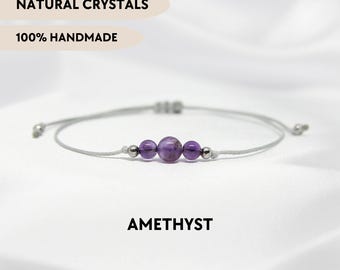 Puede incluir: Una pulsera delicada con una cuenta central de amatista morada flanqueada por dos cuentas de amatista más pequeñas y dos cuentas espaciadoras metálicas plateadas en un cordón gris. Texto: NATURAL CRYSTALS, 100% HANDMADE, AMETHYST, YPCRYSTALJEWELRY.