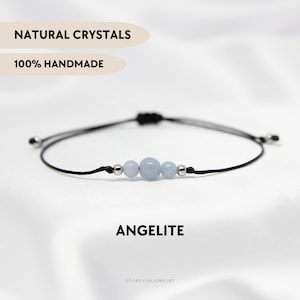 Peut inclure: Un bracelet fait à la main avec trois perles d'angélite bleu clair et des détails argentés sur un cordon noir. Le mot "ANGELITE" est affiché en dessous, avec "NATURAL CRYSTALS" et "100% HANDMADE" au-dessus.