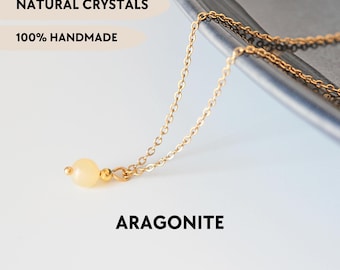 May include: A gold-toned chain necklace featuring a single, round, pale yellow aragonite bead pendant. The image includes the text NATURAL CRYSTALS, 100% HANDMADE, ARAGONITE, and YPCRYSTALJEWELRY. The necklace is displayed against a dark grey surface.