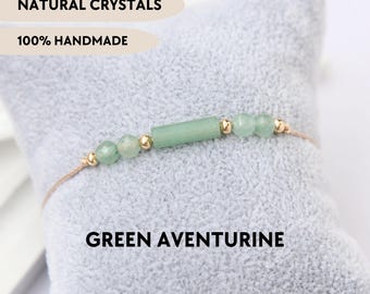 May include: A bracelet featuring a central green aventurine cylinder bead flanked by smaller round green aventurine beads and tiny gold-toned metal spacers on a thin cord. Text reads NATURAL CRYSTALS, 100% HANDMADE, and GREEN AVENTURINE.