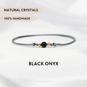 May include: A delicate bracelet featuring a single black onyx bead flanked by two small gold-toned metallic beads on a thin grey cord. The text NATURAL CRYSTALS, 100% HANDMADE, and BLACK ONYX is visible. The design includes knotted cord details.