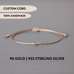 Op de afbeelding: Een dun beige koordarmband met een kleine metalen kraal in het midden. De tekst CUSTOM CORD, 100% HANDMADE en 9K GOLD | 925 STERLING SILVER is zichtbaar. De armband heeft verstelbare schuifknopen met kleine kraaltjes aan de uiteinden.