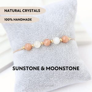 Può includere: Un braccialetto delicato con perline rotonde alternate di pietra del sole color pesca e pietra di luna bianca traslucida, accentuato da piccoli distanziatori in metallo dorato su un cordino sottile. Il testo recita NATURAL CRYSTALS, 100% HANDMADE, SUNSTONE & MOONSTONE.
