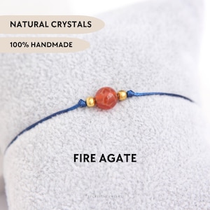 Puede incluir: Una pulsera hecha a mano con una vibrante cuenta de ágata de fuego naranja, detalles dorados y un cordón azul marino. El texto dice "NATURAL CRYSTALS", "100% HANDMADE" y "FIRE AGATE".