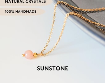 May include: A gold-toned chain necklace featuring a single round, pale peach-coloured sunstone bead accented by a small gold-toned metal bead. The image includes the text NATURAL CRYSTALS, 100% HANDMADE, SUNSTONE, and YPCRYSTALJEWELRY.